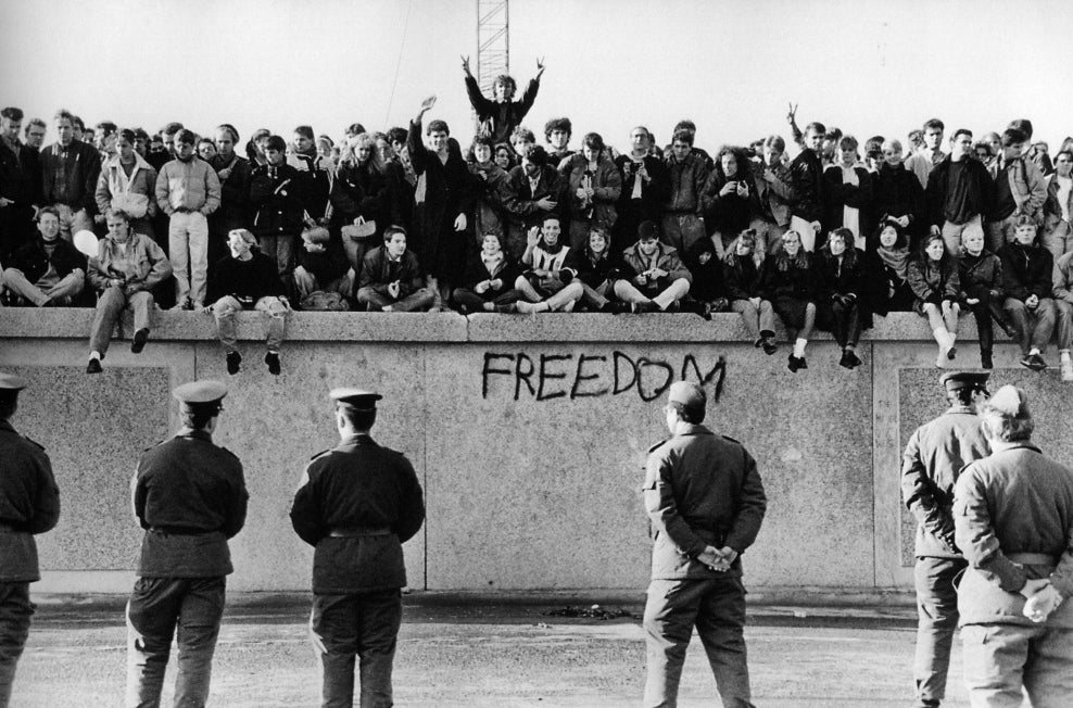 In November, 1989, East German students sit atop the Berlin Wall at the Brandenburg Gate in front of border guards.