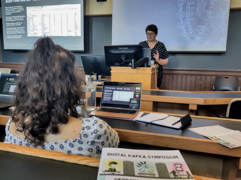 Dr. Kick presents at the podium during the Digital Kafka Symposium. The event program is visible in the foreground.