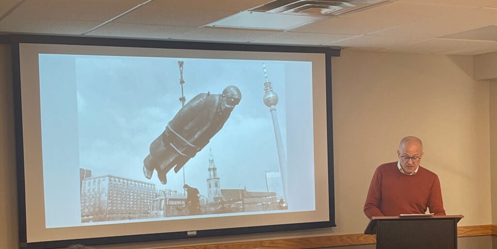 Dr. Steffen Siegel stands at a podium with an image of a statue being transported by ropes on the screen behind him